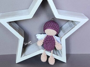 Gehäkelter Amigurumi-Engel in Flieder mit weißen Flügeln und Aufhängevorrichtung vor einem Holzstern