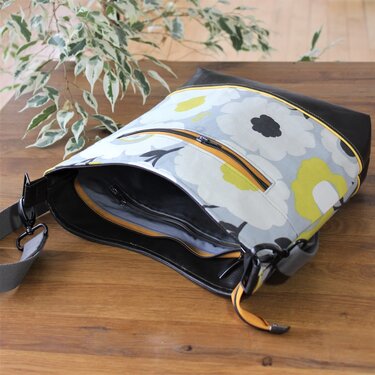 Schultertasche mit floralem Muster in Grau, Gelb und Schwarz, Kunstlederboden, orangefarbene Reißverschlüsse und verstellbarer Riemen auf Holzuntergrund.