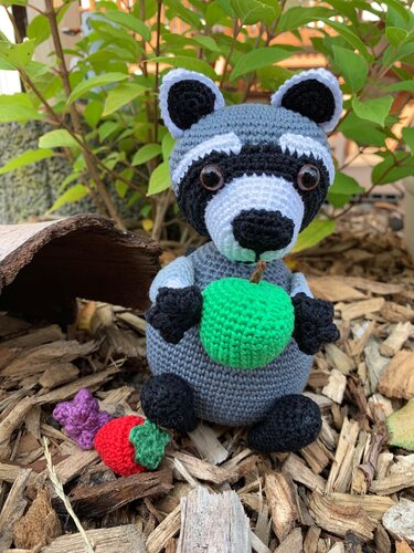Gehäkelter Amigurumi-Waschbär in Grau, Schwarz und Weiß hält einen grünen Apfel; umgeben von kleinen gehäkelten Früchten auf Holzhackschnitzeln.