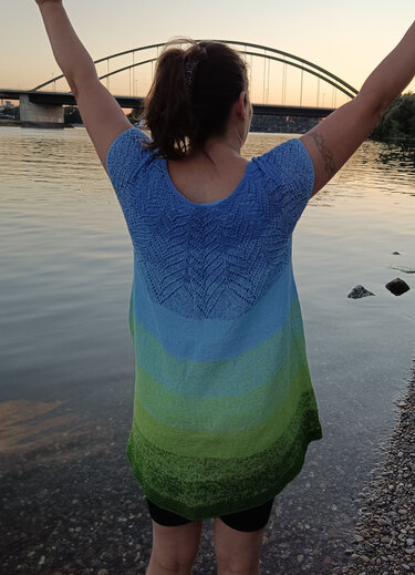 Rückenansicht einer Frau am Flussufer, sie trägt eine kurzärmelige gestrickte Tunika mit blau-grünem Farbverlauf und erhobenen Armen