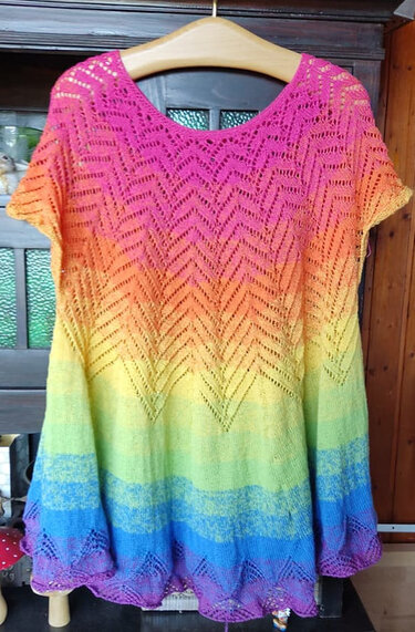 Gestricktes Kurzarmshirt in Regenbogenfarben an einem Kleiderbügel mit Zickzack‑Ajourmuster.