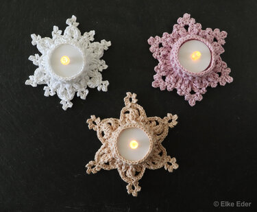 Three crocheted snowflake and star tea light holders in white, light pink, and beige, each with a lit tea light.