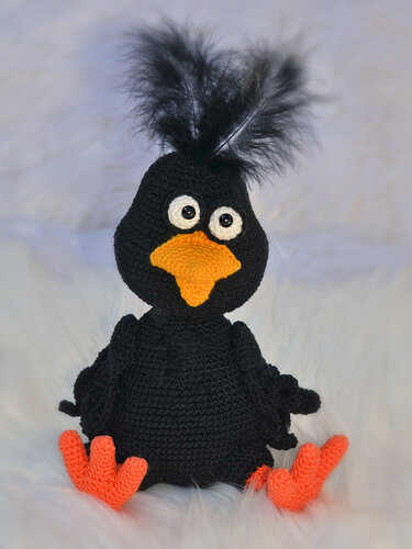 Gehäkelter schwarzer Amigurumi-Vogel mit orangefarbenem Schnabel, orangen Füßen und schwarzer Federhaube auf hellem, flauschigem Untergrund.