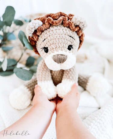Gehäkelter Amigurumi-Löwe in Beige mit brauner Mähne, gehalten von zwei Kinderhänden