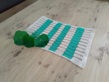 Rechteckiges gehäkeltes Deckchen mit beigen Feldern und türkisen Streifen auf hellem Holztisch, dekoriert mit drei grünen Blättern