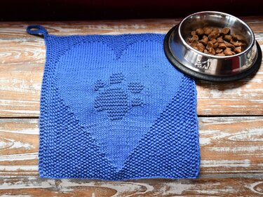 Blue knitted mat with heart and paw print beside a stainless-steel pet bowl.