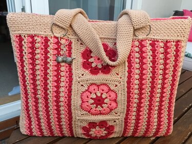 Rechteckiger gehäkelter Shopper in Beige, Rosa und Rot mit drei mittigen Granny-Square-Blumen, gestreiften Seiten, breiten Trägern, rosa Innenfutter und einem Garn-Charm