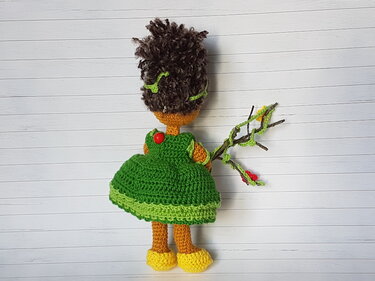 Rückseite einer gehäkelten Amigurumi-Puppe mit braunen Locken, grünem Kleid, gelben Schuhen und einem Ast mit Ranken