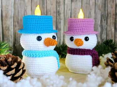 Pair of handmade crocheted snowmen with blue and purple hats and scarves, each topped with a small LED candle, surrounded by winter decor.