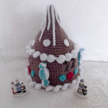 Gehäkeltes braunes Lebkuchenhaus mit weißer Dach- und Randverzierung und bunten Akzenten, dazu zwei kleine Figuren