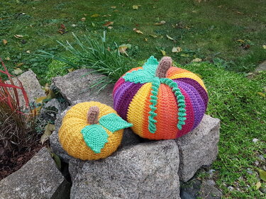 Detailansicht zweier Häkelkürbisse mit Rippenmuster, grünen Blättern, braunen Stielen und einer Spiralranke auf Steinen.