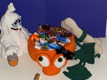Orange gehäkelte Süßigkeitenschale mit großen Augen und schwarzer Spinne, gefüllt mit Süßigkeiten; daneben ein weißer Häkelgeist und grüne Häkel-Accessoires