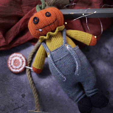 Knitted pumpkin amigurumi doll with grey overalls holding a striped lollipop