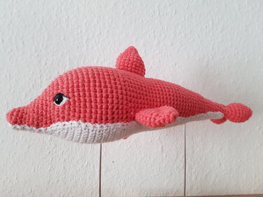 Korallenrotes gehäkeltes Delfin-Amigurumi mit weißem Bauch und schwarzem Auge vor weißer Wand