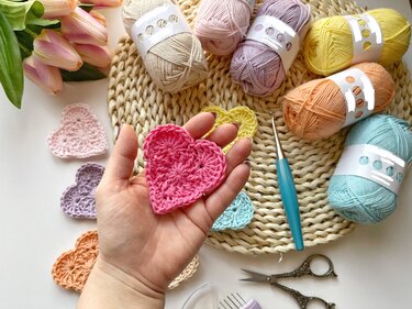 Hand holding a small pink crochet heart with pastel yarns and a crochet hook