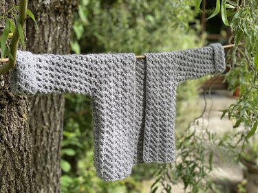 Gray crocheted open-front jacket hanging on a wooden rod between trees, showing textured stitch detail
