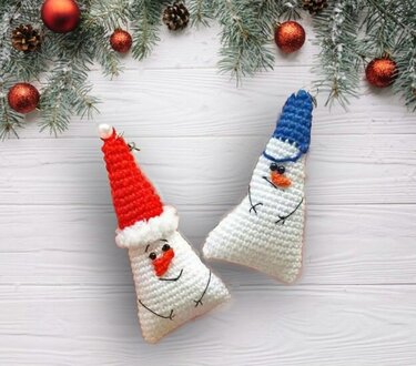 Zwei dreieckige gehäkelte Schneemann-Schlüsselanhänger mit roter und blauer Mütze, gesticktem Gesicht und Aufhängeschlaufe auf hellem Holzhintergrund