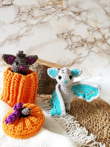 Zwei gehäkelte Amigurumi-Fledermäuse, eine grau-türkis mit ausgebreiteten Flügeln, eine dunkelgrau in einem orangefarbenen Kürbissäckchen mit Deckel