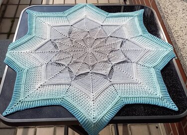 Star-shaped crocheted doily with gray-to-teal ombre on a dark table.