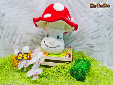 Fröhlicher Häkelpilz mit rotem Hut, stehendes Amigurumi in Holzkiste neben weißen Orchideen und Dekoschmetterling