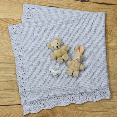 Hellgraue gestrickte Babydecke mit Spitzenrand, darauf ein Teddybär, ein Hase und ein Schnuller auf Holzboden.