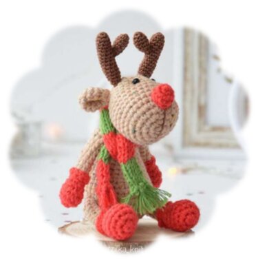 Sitzendes gehäkeltes Rentier-Amigurumi mit roter Nase, grünem Schal mit Herzmotiven und roten Handschuhen vor hellem Hintergrund.