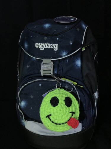 Gehäkelter leuchtgrüner Smiley-Anhänger mit roter Zunge an dunkelblauem Rucksack mit Sternenmuster.