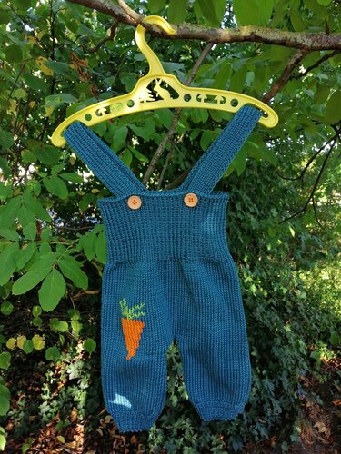 Petrolfarbene gestrickte Baby-Latzhose mit Karotten-Applikation und Holzknöpfen an gelbem Kleiderbügel