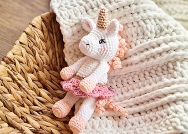 Handmade white crochet unicorn amigurumi with pink skirt and gold horn resting in a basket on a chunky knit blanket.