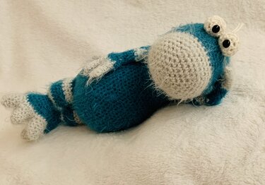Blau-creme gehäkeltes Amigurumi-Fantasiewesen liegend mit großer runder Schnauze und Kulleraugen