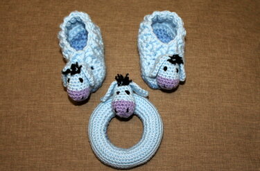 Hellblaue gehäkelte Babyschuhe mit Eselköpfen und passender Ringrassel auf braunem Stoff