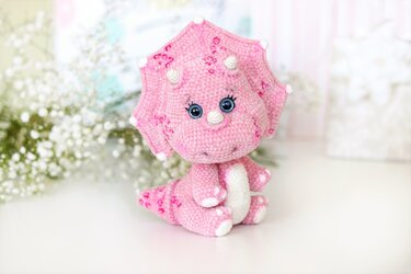 Pink crocheted triceratops plush with blue safety eyes, white horns, and a frilled collar.