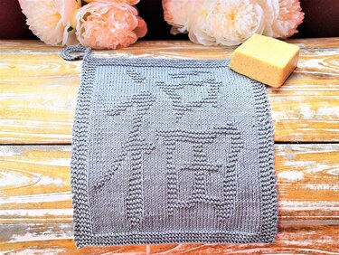 Light blue-gray knitted potholder with raised character motif and corner loop on wooden surface with flowers and soap.