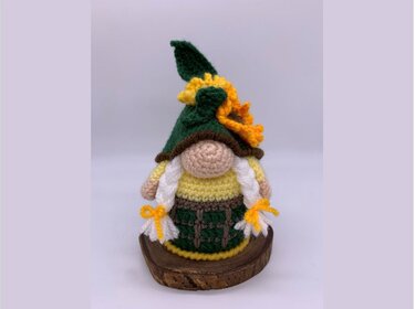 Crocheted amigurumi gnome with dark green pointed hat, sunflower accent, yellow body, green skirt and white braided pigtails on a wood slice.