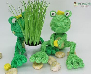Zwei gehäkelte grüne Amigurumi-Frösche, einer hält einen weißen Topf mit Gras, auf hellem Hintergrund mit Kieselsteinen.