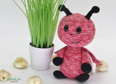 Rosa gehäkeltes Amigurumi-Käferchen mit schwarzen Antennen neben einem kleinen Topf mit Gras