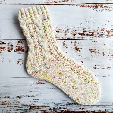 Gestrickte cremefarbene Socke mit Zopfmuster, Ajourlöchern und bunten Sprenkeln auf hellem Holzuntergrund