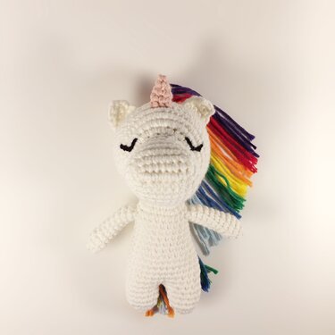 Weißes gehäkeltes Amigurumi-Einhorn mit rosa Horn, geschlossenen Augen und bunter Regenbogenmähne auf hellem Hintergrund.