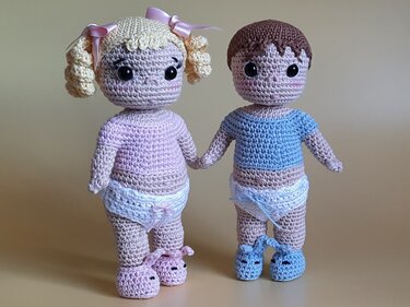 Zwei gehäkelte Amigurumi-Puppen, Hand in Hand, mit großen schwarzen Augen, Locken mit Schleifen und kurzem braunem Haar, Windelhöschen und Häkel-Schühchen