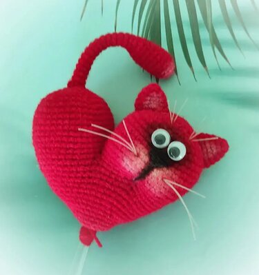 Rote, herzförmige Amigurumi-Katze mit Wackelaugen auf pastellgrünem Untergrund