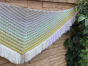 Gehäkeltes Dreieckstuch in Pastellfarben (Grau, Grün, Gelb) mit Lochmuster und weißen Fransen, an einer Mauer befestigt.
