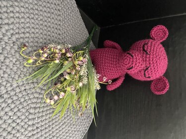 Pinkes gehäkeltes Amigurumi-Mäuschen mit geschlossenen Augen neben kleinem Blumenstrauß auf grauem Kissen