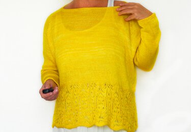 Nahaufnahme eines gelben gestrickten Pullovers mit Lochmuster am unteren Saum und weitem U-Boot-Ausschnitt. Eine Hand hält ein kleines schwarzes Objekt.