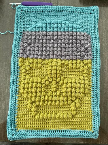 Rectangular crocheted panel with a raised bobble-stitch skull motif in yellow and gray, framed by a turquoise border and a purple-handled crochet hook at the top