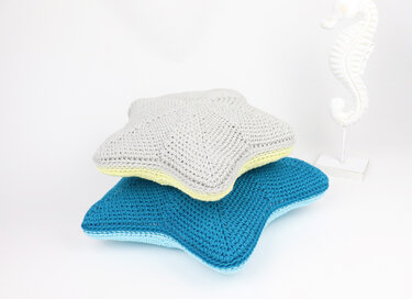 Two crocheted starfish pillows, one grey and one blue, stacked on a white surface next to a white seahorse decoration.