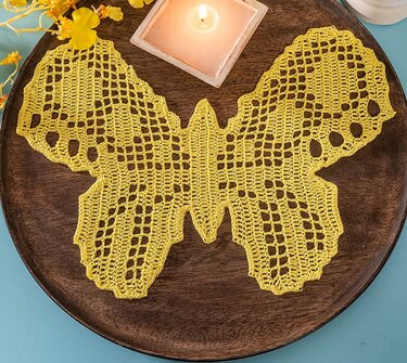 Gelbes gehäkeltes Schmetterlingsdeckchen auf dunklem Holztablett mit Kerze und gelben Blüten.