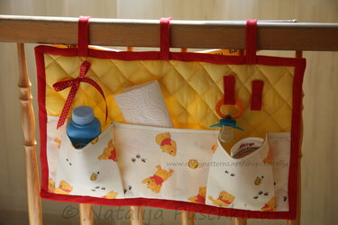 Gestepptes Hänge-Utensilo in Gelb und Rot mit Bärenmotiv-Taschen, gefüllt mit Babyflasche, Schnuller, Papiertuch und Creme, hängend an einem Holzgeländer