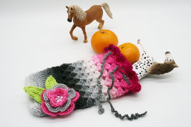 Gehäkelter Beutel in Grau‑ und Pinktönen mit mehrlagiger Blüte und Blättern, mit Zugband, liegend neben Orangen und Spielzeugpferden auf weißem Hintergrund.