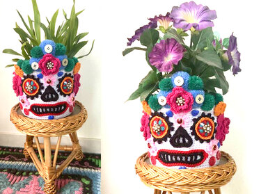 Bunter gehäkelter Übertopf in Totenkopfform mit gehäkelten Blumen und einer Topfpflanze auf einem Rattanhocker.