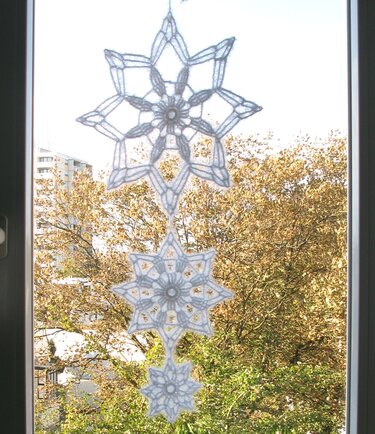 Drei weiße gehäkelte Schneeflocken als Kette hängend vor einem Fenster mit Bäumen im Hintergrund
